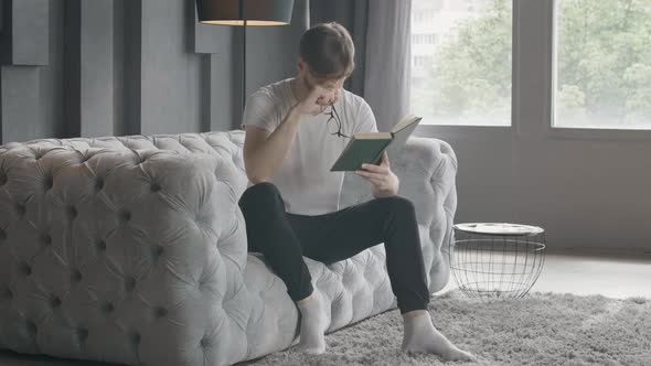 Brunette Man Taking Off Eyeglasses and Rubbing Eyes As Reading Book at Home. Wide Shot Portrait of alt