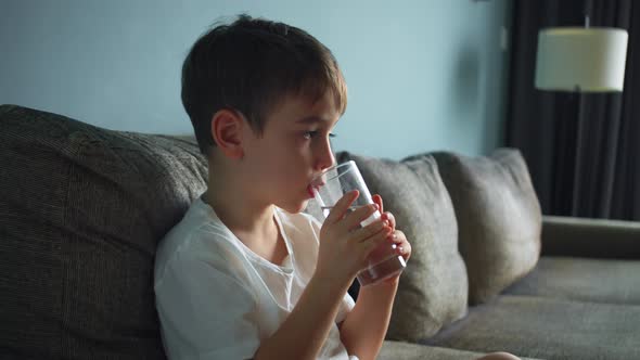 Child is Drinking a Glass of Water Sitting on Sofa at Home While Watching TV alt