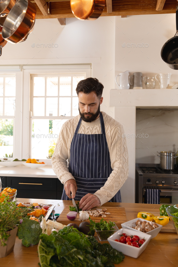 Caucasian man preparing food, concentrating on chopping vegetables, wearing apron in kitchen