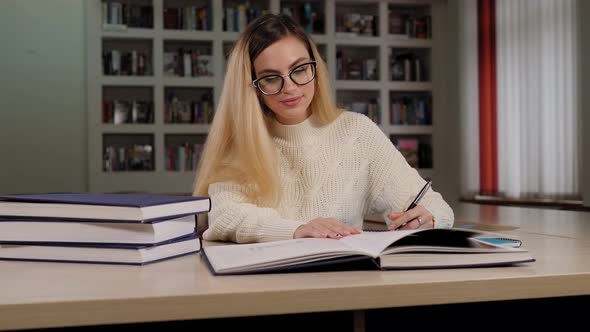 A Student Girl with Glasses Is Sitting at a Library Table with Books and Writing alt