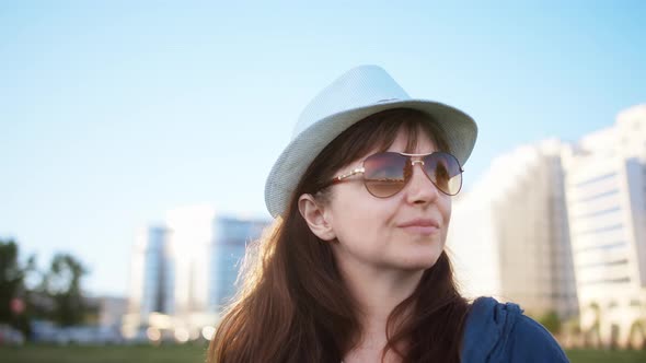 Happy Brunette Woman in a Hat Stands Against the Background of Houses and Looks Around