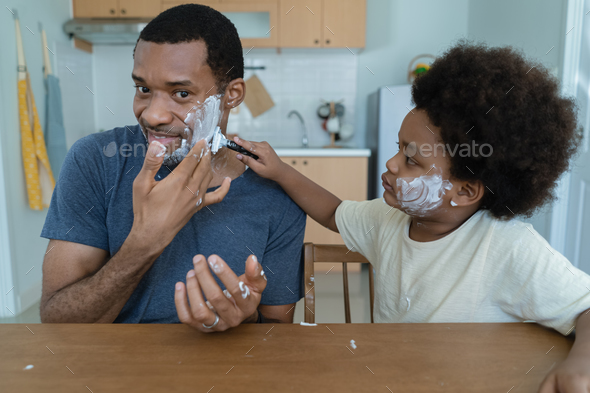 African American Father and son having fun while shaving and looking at mirror with kid razor help