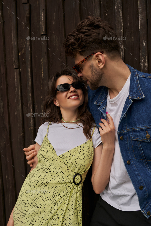 Positive brunette woman in stylish sundress and sunglasses touching bearded boyfriend in denim vest