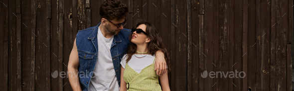 Brunette man in sunglasses and denim vest hugging joyful girlfriend in sundress and standing