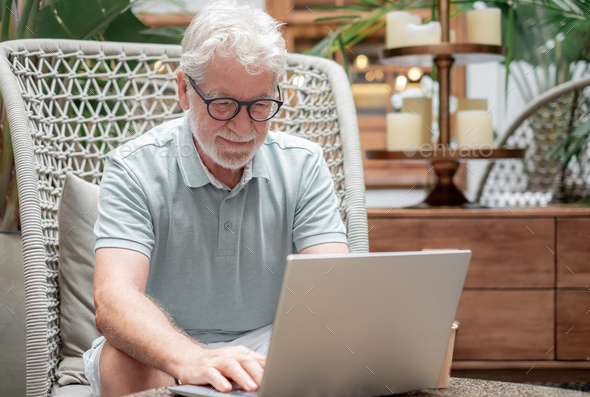 Senior bearded man sitting indoor using laptop typing on keyboard while surfing the net