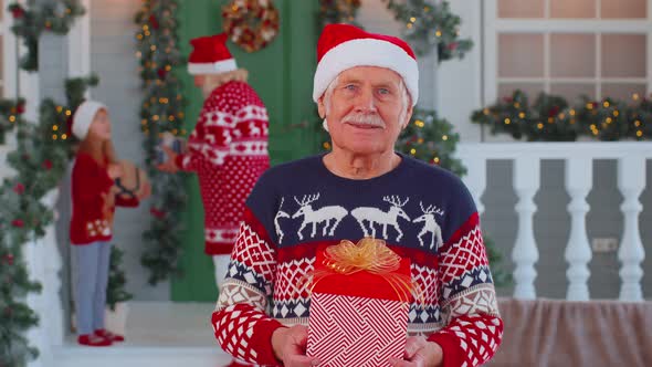 Portrait of Grandfather Man Presenting Gift Box Smiling Near Decorated Christmas House with Family alt