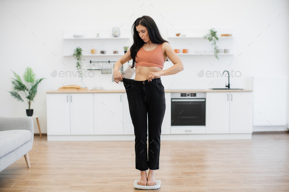 Amazed female in loose pants standing on scales in kitchen