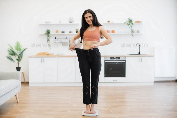 Amazed female in loose pants standing on scales in kitchen