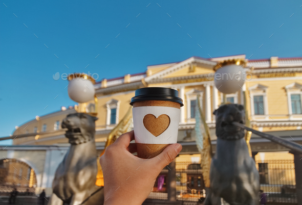 cup of coffee with carved white heart in woman's palm on background of bank bridge in St. Petersburg