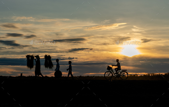 Silhouette of woman carry pack of rice and walk along the road near water reservoir follow by child
