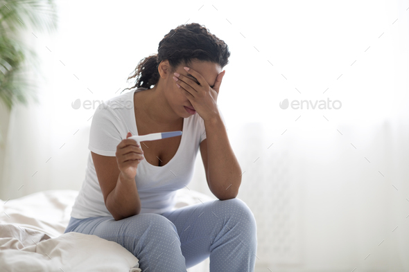 Unwanted Pregnancy Concept. Desperate black woman holding pregnancy test, sitting on bed