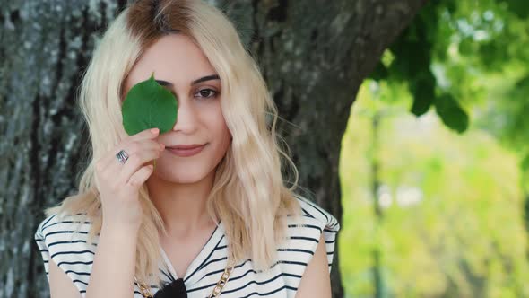 Blond Caucasian Girl Holding a Small Green Leaf Close to the Eye alt
