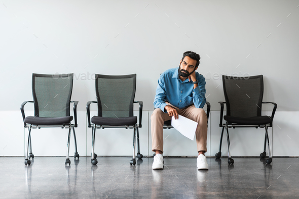 Bored indian man with CV tired of waiting for employment interview at office lobby, sitting on chair