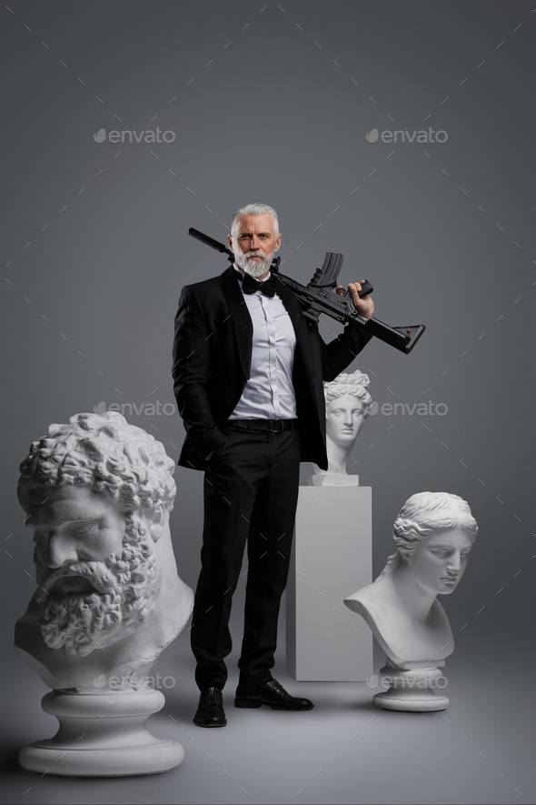 Stylish older man holding a gun stands surrounded by three ancient sculptures