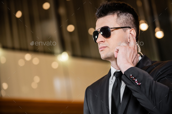 handsome bodyguard in sunglasses, handsome man in suit and tie touching  earpiece in lobby of hotel, security, career, communication, vigilance,  privat Stock Photo - Alamy