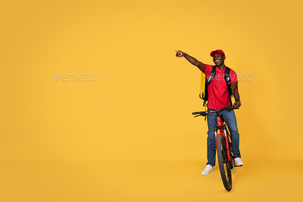 Happy young african american delivery man in uniform with backpack rides bike, points finger to free