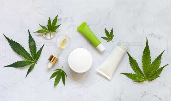 Pipette with oil, cream jars and cosmetic tubes near green cannabis leaves on white table top view