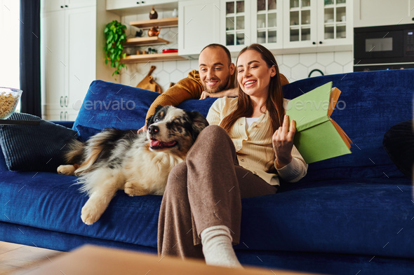 Smiling couple with book relaxing near border collie on couch and popcorn in living room at home