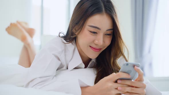 Asian attractive woman lying down on bed, use mobile phone chat with friend at home in morning. alt