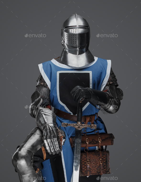 Medieval knight in armor stands leaning on his sword, against a gray
