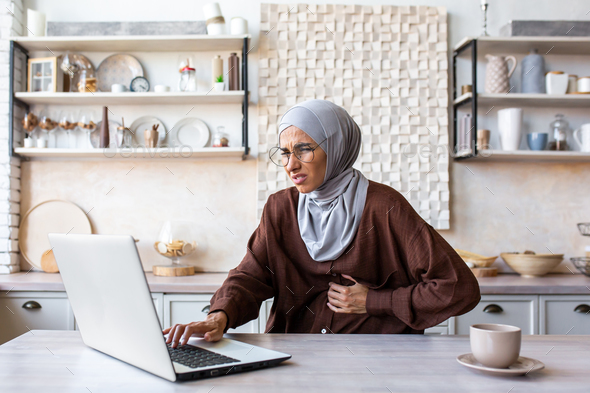 Young Muslim woman in hijab working, studying at home using laptop. Holds his stomach with his hand
