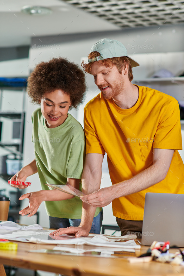 Smiling craftsman holding printing layer near african american colleague, clothes and laptop