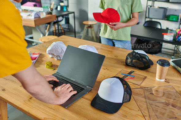 Cropped view of young craftsman using laptop near coffee to go and african american colleague