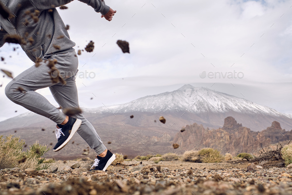 Crop man in activewear running on dirty terrain near mountains