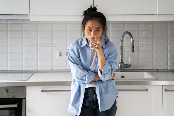 Unhappy disheveled Asian girl drinks water while standing in kitchen waking up late morning bad mood