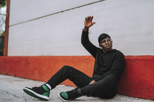 Sporty Black man in black sweaters and jogging pants sitting on concrete pavement outside a building