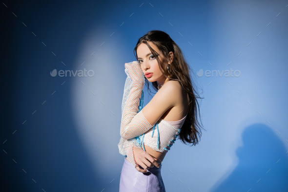 Young and trendy asian model in skirt and mesh top looking at camera on blue background with shadow