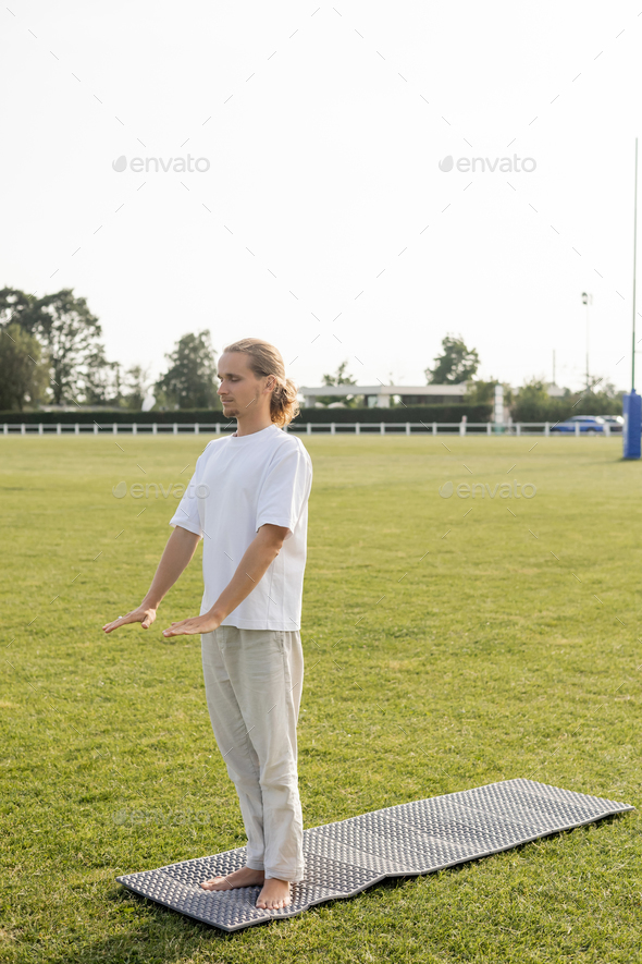full length of long haired man in linen pants and white t-shirt meditating with closed eyes