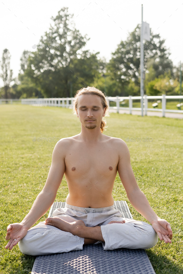 shirtless man in linen pants meditating in lotus pose while sitting on yoga mat with closed eyes