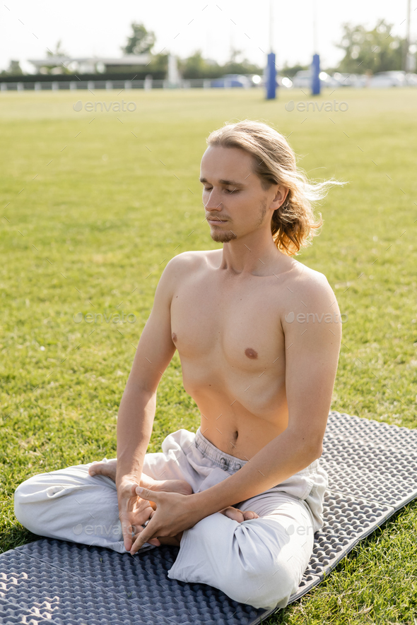 shirtless long haired man in linen pants meditating in lotus pose with closed eyes on grassy
