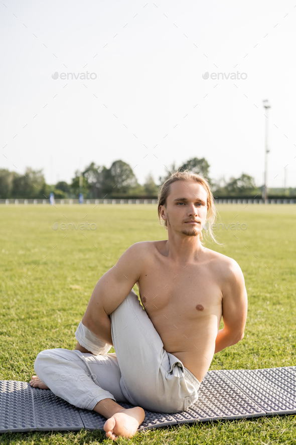 barefoot shirtless man in linen pants looking away while practicing yoga in sage pose on green