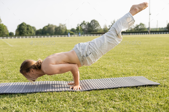 side view of young shirtless man in linen pants meditating in peacock pose on yoga mat on green