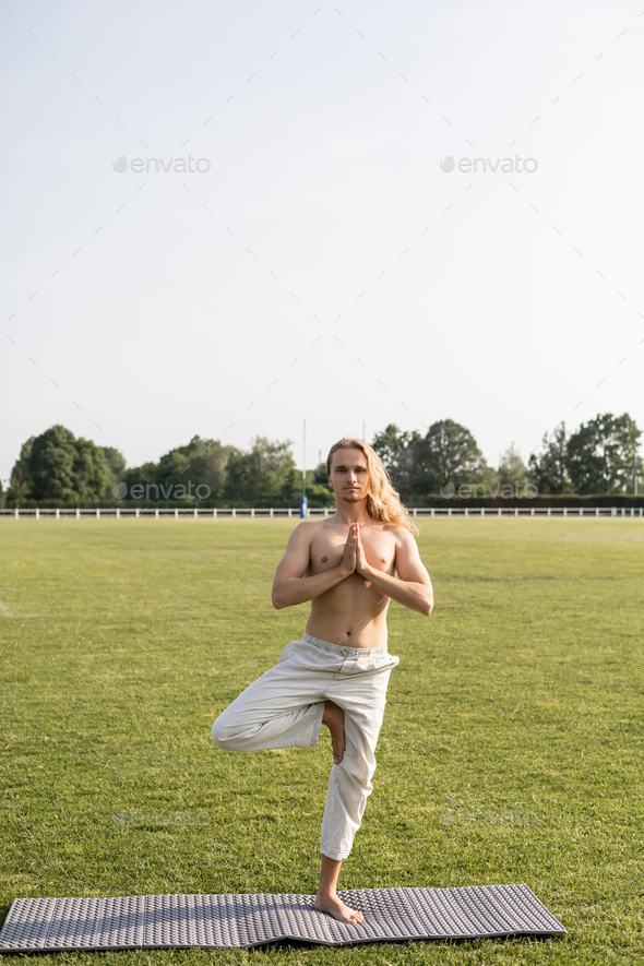 full length of shirtless man in linen pants standing in tree pose with anjali mudra gesture