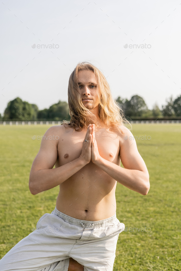 long haired and shirtless man in linen pants meditating in tree pose with praying hands on green