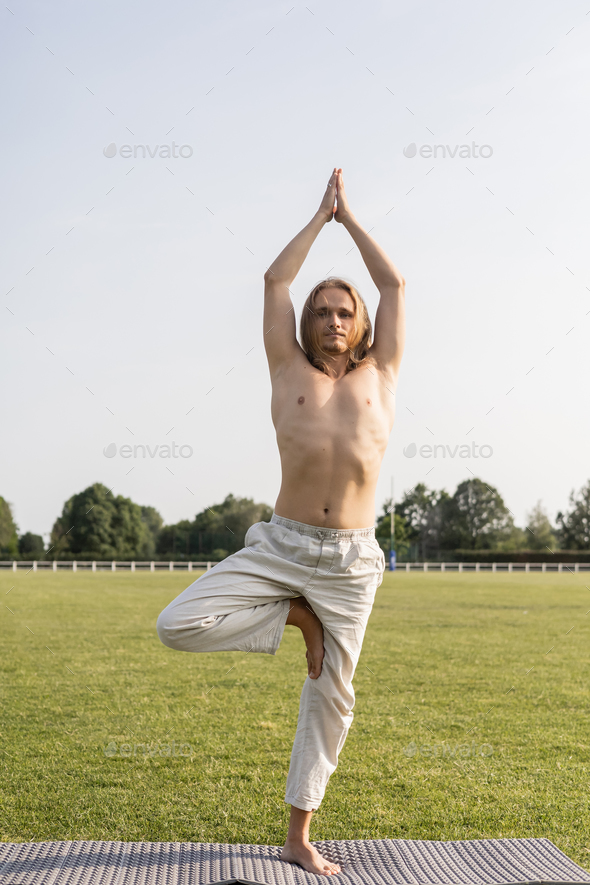 shirtless long haired man in linen pants meditating in tree pose with raised praying hands on yoga