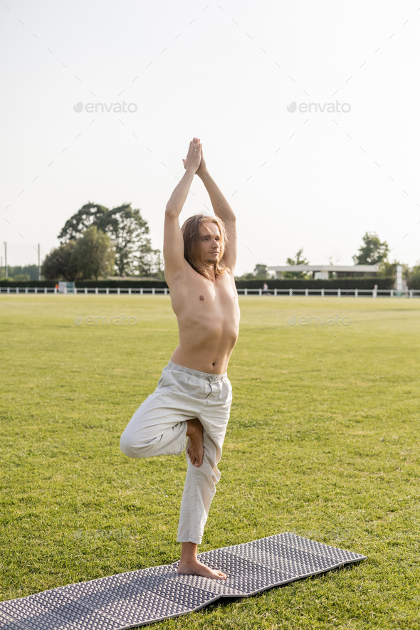shirtless barefoot man in linen pants meditating in tree pose with raised hands on yoga mat