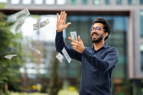 Overjoyed rich young indian businessman throwing money dollar banknotes