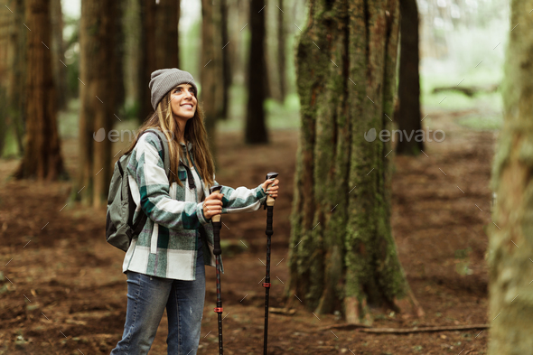 Happy young caucasian woman in jacket with trekking sticks walk enjoy trip alone in forest