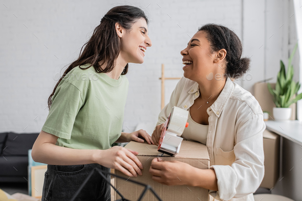 joyful multiracial woman holding tape dispenser near carton box and happy lesbian partner