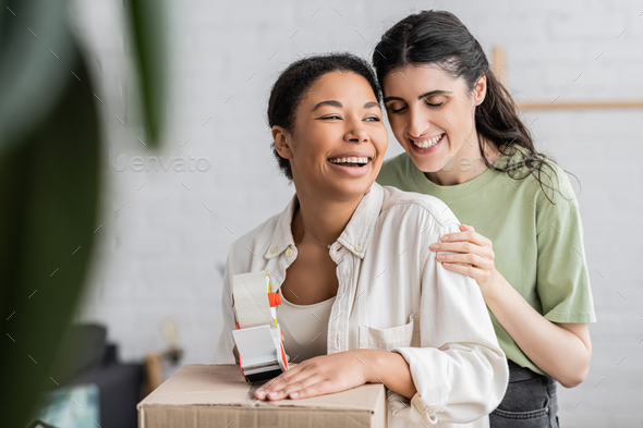 cheerful multiracial woman holding tape dispenser near carton box and laughing with lesbian partner