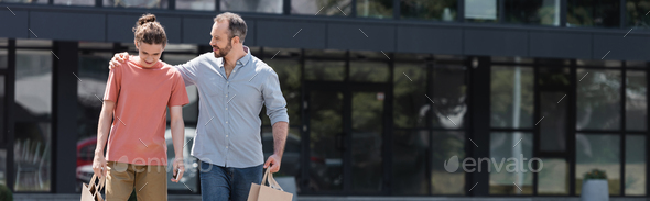 cheerful dad hugging teenage son while walking from mall with shopping bags, banner