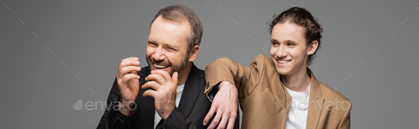 cheerful teenage boy and overjoyed dad in suits laughing isolated on grey, banner