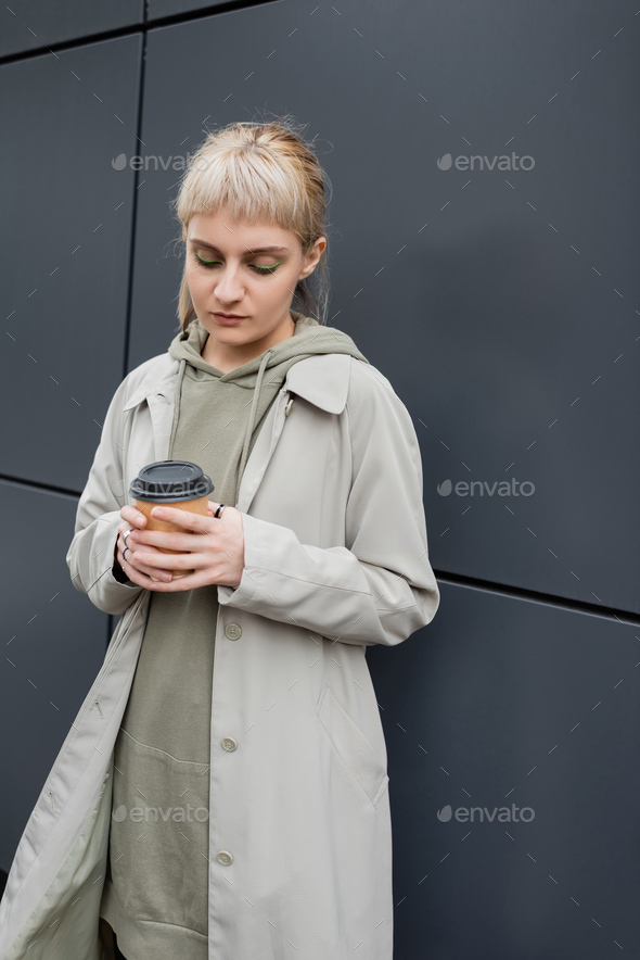 young woman with blonde hair with bangs standing in coat and hoodie while holding paper cup