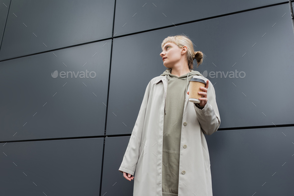low angle view of stylish young woman with blonde hair with bangs standing in coat and hoodie