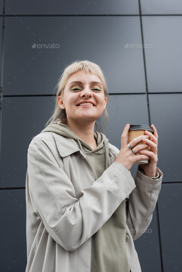 low angle view of happy young woman with blonde hair with bangs standing in coat and hoodie