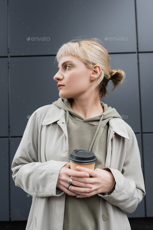 stylish young woman with blonde hair with bangs standing in coat and hoodie while holding paper cup
